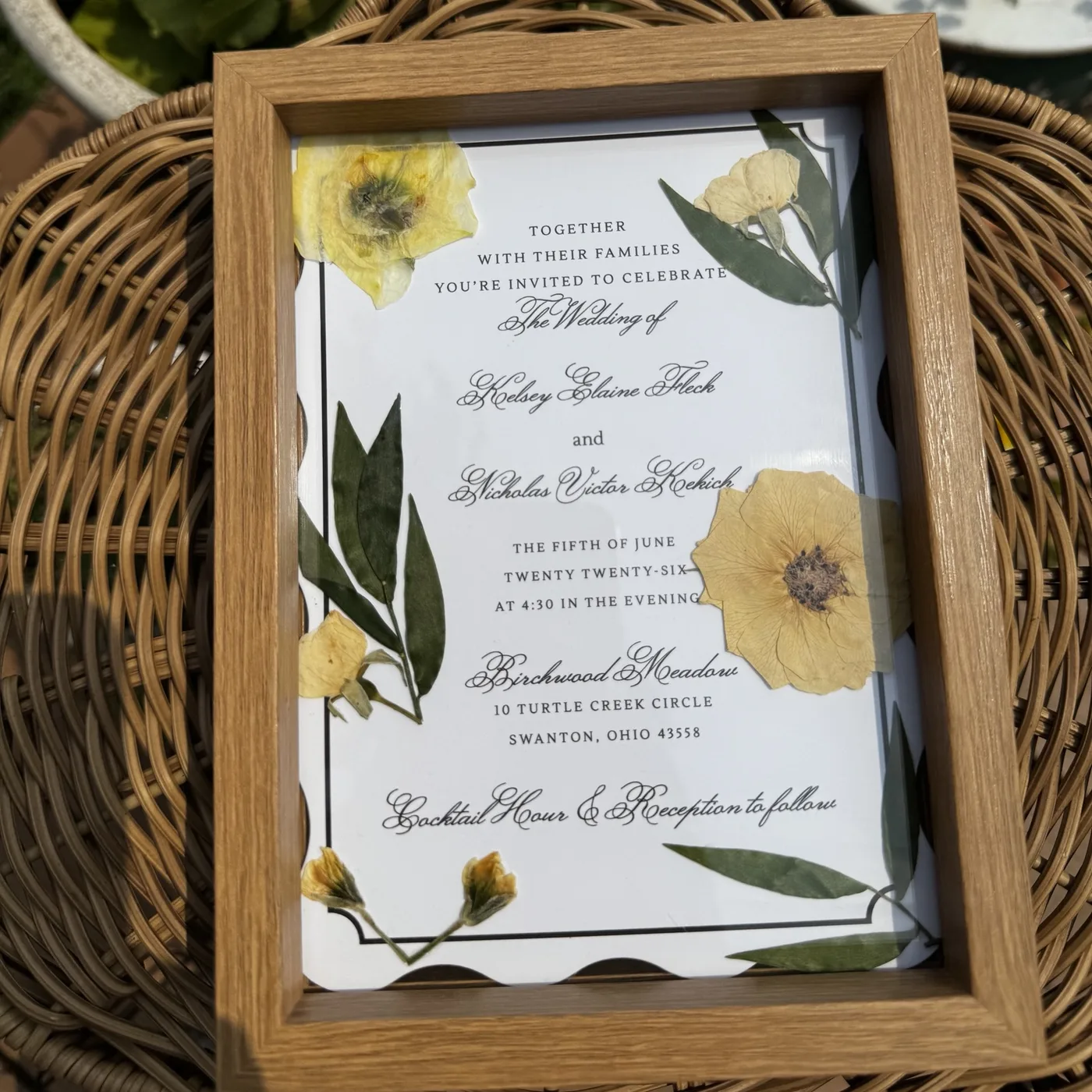 A framed wedding invitation bordered with pressed yellow flowers and greenery
